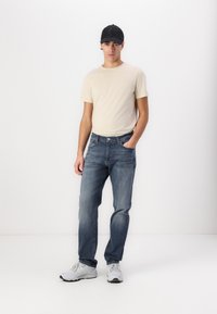T-shirt beige clair, jeans en denim bleu, baskets grises, casquette noire. Design slim fit, détails minimaux, texture lisse, style décontracté.