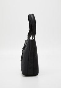 Sac cabas rectangulaire noir avec deux anses et une poche latérale zippée, présenté de profil sur un fond clair uni.