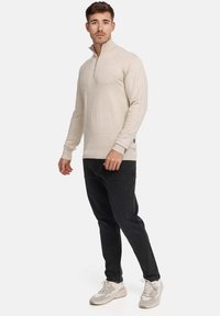 Maglione beige con zip, maniche lunghe, polsini e orlo a coste, abbinato a jeans neri e sneakers grigio chiaro. Tessuto semplice e strutturato.
