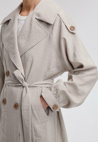 Hele beež trench coat, mis on valmistatud tekstuurilisest kangast, koos topelt-nööbiga disaini, laia krae ja suurte, läbipaistvate nuppudega.