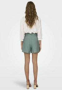 Světle zelené šortky na míru s vysokým pasem a poutky na opasek, zkombinované s bílou blůzou, pohled zezadu. Hladká tkanina a klasický design.