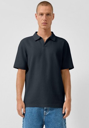 Man met kort haar en oorbellen, die een donker poloshirt met korte mouwen en een blauwe spijkerbroek draagt, staand tegen een effen lichte achtergrond.