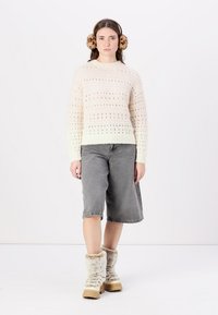 Strikkegenser i krem med åpenmønster, parret med grå denim culottebukser. Bruker beige støvler med pelskant og ørevarmere med dyremønster.