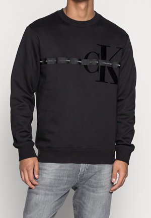 Hombre vistiendo un sudadera negra de Calvin Klein con una franja horizontal brillante y un gran logo "cK", combinado con jeans gris claro.