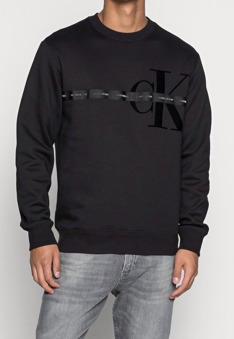 Uomo che indossa un felpa nera Calvin Klein con una striscia orizzontale lucida e un grande logo "cK", abbinata a jeans grigi chiari.