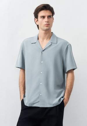 Jeune homme les mains dans les poches, portant une chemise grise claire à manches courtes boutonnée et un pantalon noir, regardant légèrement sur le côté.