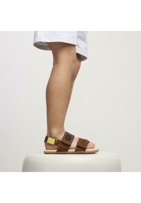 Sandali marroni con cinturini regolabili, un accento giallo e un logo circolare. Suola piatta e design aperto, adatti per il clima caldo.