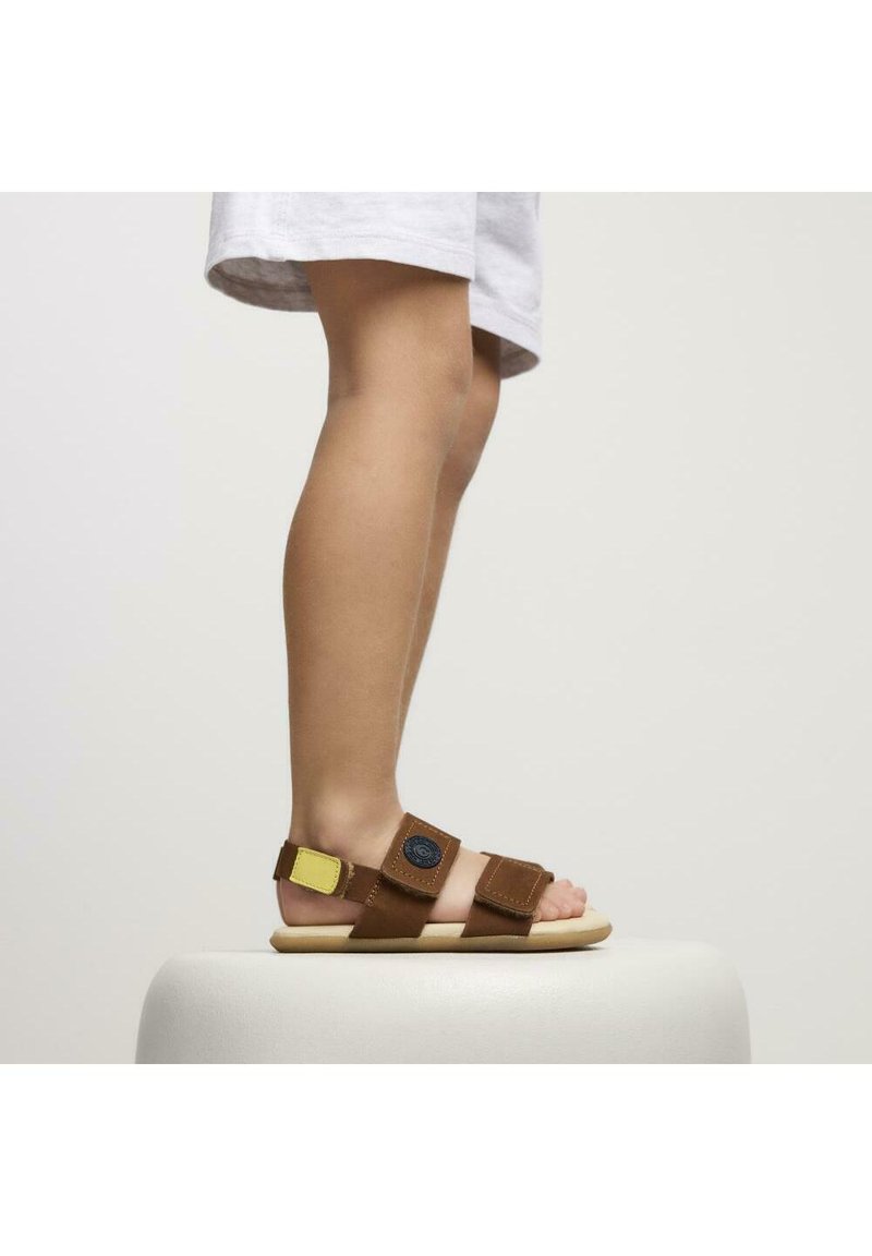 Sandali marroni con cinturini regolabili, un accento giallo e un logo circolare. Suola piatta e design aperto, adatti per il clima caldo.