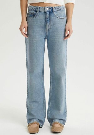 Proste, jasnoniebieskie jeansy z denimu noszone z beżowymi balerinami, pokazane od talii do stóp na jednolitym tle.