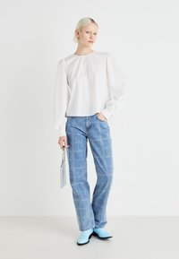 Blouse blanche à manches longues et bouffantes, associée à un jean en denim bleu clair à clous et des bottines bleu ciel, tenant une pochette argentée.