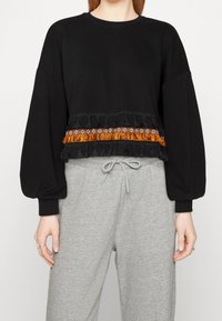 Sweat-shirt noir court avec manches longues bouffantes, doté d'un bord orange à franges et de perles délicates à l'ourlet. Associé à un pantalon de survêtement gris.