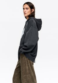 Oversizowa ciemnoszara bluza z kapturem z grafiką, z troczkiem przy kapturze i ściągaczami na rękawach, zestawiona z oliwkowymi spodniami o fakturowanej powierzchni.