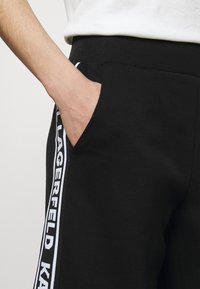 Czarne dresy o gładkiej fakturze, z boczną kieszenią i białym paskiem z logo wzdłuż nogawki. W zestawie elastyczny pasek w talii.