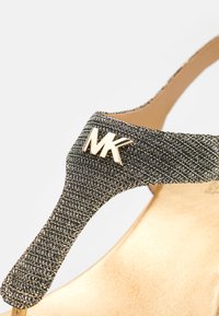 Gros plan sur une bride de sandale noire texturée avec un détail logo "MK" doré au-dessus d'une semelle beige.
