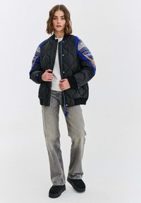 Prešívaná čierna bomber bunda s modrými akcentmi a vzorovanými rukávmi, so zapínaním na zips, rebrovanými manžetami a dekoratívnym strapcom.