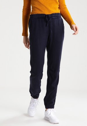 Personne portant un pantalon de jogging bleu marine à cordon, des baskets blanches et un haut côtelé à manches longues moutarde, debout devant un fond uni.