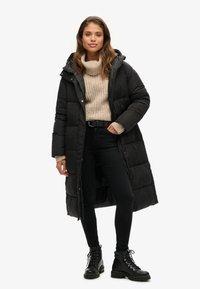 Sort pufferjakke med hætte, der har et langt design og quiltet tekstur, lagdelt over en lys beige strikket sweater og sorte stramme jeans.