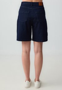 Shorts di jeans di colore blu scuro, con un taglio classico, tasche posteriori e una patch in pelle marrone in vita. Texture liscia, design casual.