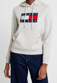 Sudadera ligera color gris claro con un bolsillo canguro en el frente, capucha con cordón y un logo bordado en azul marino, blanco y rojo en el pecho.