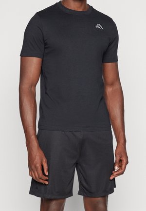 Mann trägt ein schwarzes T-Shirt mit kurzen Ärmeln und schwarze Sportshorts und steht vor einem schlichten hellen Hintergrund.