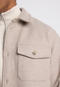 Beige villatakki, jossa on kaulus, vetoketjukiinnitys ja rintatasku, jossa on nappi. Pehmeä pinta ja hieman pörröinen viimeistely.