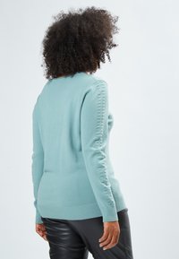 Pullover lungo verde menta con maniche lunghe, orlo a coste e dettagli laterali; materiale morbido, design aderente e piccoli bottoni al collo.