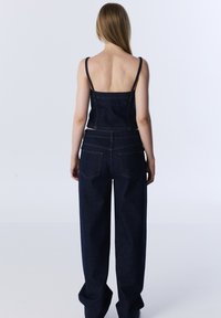 Salopette en denim foncé avec bretelles, corsage ajusté et jambes larges avec poches arrière. Texture lisse et accents de quincaillerie minimaux.