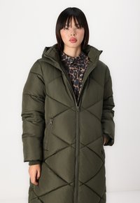 Manteau matelassé vert olive avec capuche, doté d'un design en diamant, fermeture éclair à l'avant et poches latérales. Texture lisse et matériau léger.