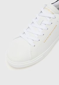 Sneaker en cuir blanc avec des lacets plats blancs, branding doré "Tommy Hilfiger", bout rond, et texture lisse sur la tige et la semelle.