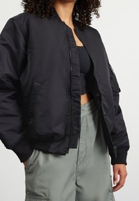 Zwarte bomberjack met een ritssluiting, geribde manchetten en een zijzakdetail. Gemaakt van een glad materiaal met een licht glanzende afwerking.