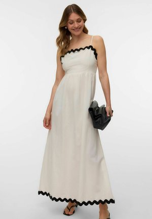 Kobieta w białej sukni maxi z czarną falbanką, trzyma czarną kopertówkę i stoi na jednolitym białym tle.