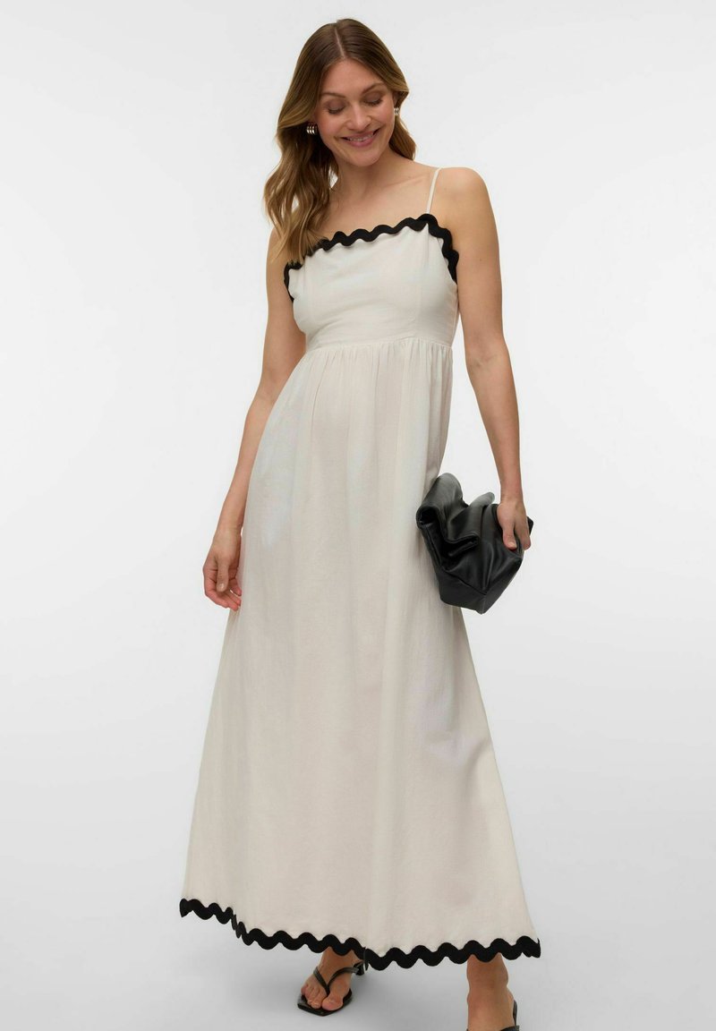 Femme portant une robe maxi blanche avec une bordure festonnée noire, tenant une pochette noire, et se tenant devant un fond blanc uni.