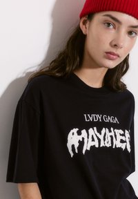 Musta t-paita, jossa on ylipitkä malli, ja jossa on valkoista tekstiä "LVDCY GAGA MAYHEM" paksussa, vääristyneessä fontissa. Liukas kankaan pinta.