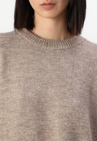 Beige stickad tröja med ribbad rundhals, mjuk textur och ett subtilt melerat mönster. Närbild på tyget.