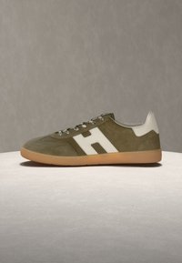 Baskets en daim vert olive avec languette de talon blanche, logo "H" blanc sur le côté, lacets à motifs et semelle en caoutchouc naturel sur surface claire.