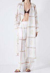 Weiß bestickter Kimono mit floralen Mustern, kombiniert mit weißen lockeren Hosen und einem braunen Oberteil, das zarte Spitze und weite Ärmel hat.