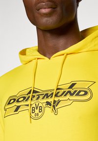 Gul hættebluse med en sort grafisk design med "DORTMUND" og BVB-logo. Tekstureret stof med justerbare snore i hætten.