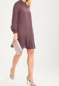 Långärmad, knälång klänning i mörkröd med en strukturerad mönster, med en volangkant. Kombinerad med en ljusgrå kuvertväska och klackar.