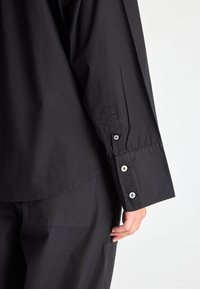 Černá košile s dlouhým rukávem z bavlny, s knoflíkovým manžetou a volným střihem. Má zaoblený spodní lem a jemnou texturu.