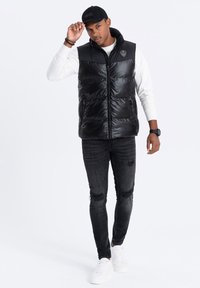 Schwarze Steppweste mit zwei seitlichen Reißverschlüssen und Logo, getragen über einem weißen Langarmshirt und kombiniert mit ausgewaschenen schwarzen Jeans und weißen Turnschuhen.