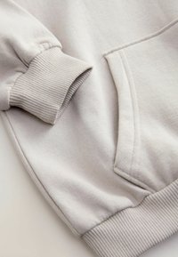 Manche de sweat-shirt beige clair et poche avant avec poignets et ceinture côtelés, montrées en gros plan sur un fond neutre.