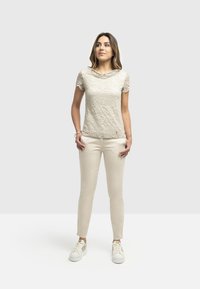 Top in pizzo beige con maniche corte e scollatura a drappo, abbinato a pantaloni a sigaretta beige chiaro e sneakers bianche. Design semplice e texturizzato.