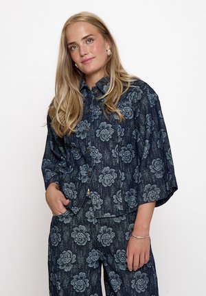 Blondine met een donkerblauwe, dichtgeknoopte jas en bijpassende broek met een groot lichtblauw bloemenpatroon, staand met één hand in de zak.