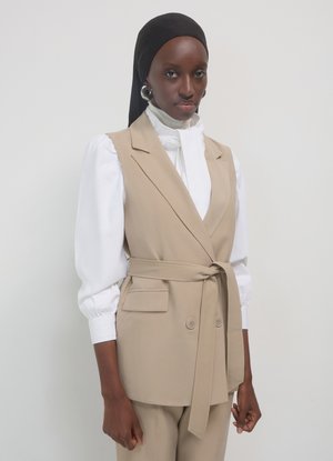 Gilet beige su misura con vita annodata, due tasche frontali e chiusura con doppio bottone, indossato sopra una camicetta bianca con maniche a sbuffo.