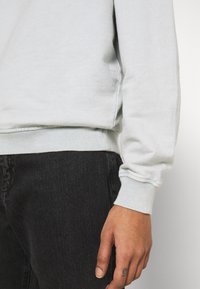Felpa grigia chiara con orlo e polsini a costine, realizzata in materiale morbido. Posizionata accanto a jeans neri, mostrando trama e vestibilità.