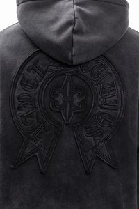 Μαύρο φούτερ με κουκούλα που διαθέτει έντονη ανάγλυφη σχεδίαση ενός κυκλικού έμβληματος με κορδέλες, κατασκευασμένο από απαλό, υφασμάτινο υλικό.
