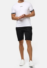T-shirt blanche à manches courtes avec des accents noirs sur les manches, assortie à un short noir et des baskets blanches. Présente un logo simple sur le t-shirt.