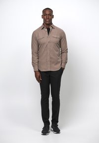 Lichtbruine, knoopsluiting shirt met twee borstzakken. Gecombineerd met zwarte broek en zwarte sportschoenen. Model staat tegen een effen achtergrond.