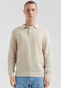 Beige strukturierte langärmlige Poloshirt mit kragenförmigem Ausschnitt und drei Knöpfen, mit gerippten Bündchen und Saum. Kombiniert mit hellblauen Jeans.