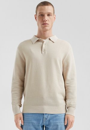 Beige textured long-sleeve polo shirt with a collared neckline and three buttons, featuring ribbed cuffs and hem. Worn with light blue jeans.